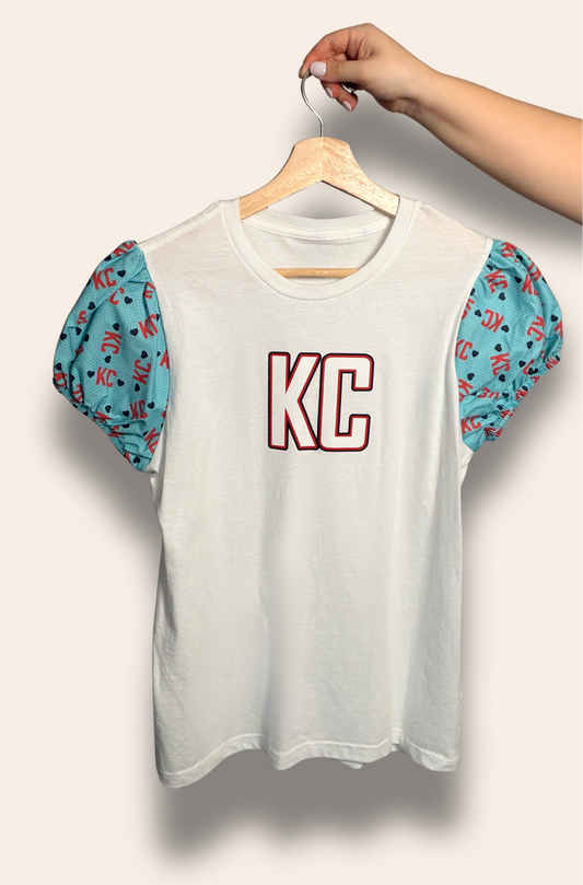 For the girls who cheer loud and dress cute doing it.

Our KC Current Sweetheart Puff Tee features a classic white base with bold KC lettering and dreamy teal puff sleeves covered in playful KC and heart details. It’s the perfect mix of feminine and fierce, made for match days, summer nights, and showing your Current pride in style.

Soft, flattering, and made to stand out in a sea of teal.
