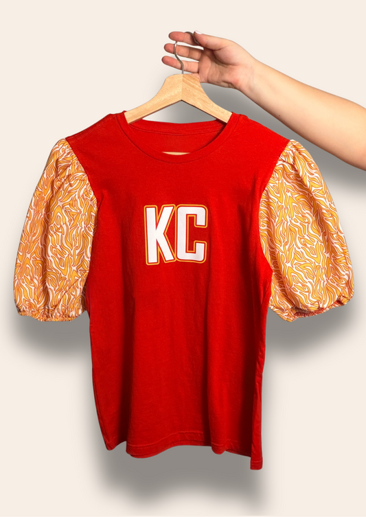 For the girls who bring electric energy to the Kingdom.

Our KC Kingdom Electric Puff Tee features a bold red base with vibrant gold statement puff sleeves inspired by the iconic Chiefs colors. Feminine, fierce, and made to stand out, it’s the perfect look for tailgates, touchdowns, and cheering on Kansas City in style.

Comfortable, flattering, and made for your game day era.
